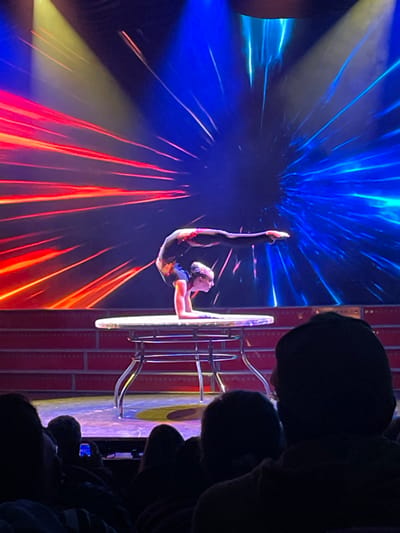 A performer balances upside down with legs extended upward, gripping a large, silver hoop supported by a metal stand. The background features dynamic, radiating lines of blue and red light, creating a vibrant visual effect. The performer wears a dark bodysuit with shimmering accents. Silhouetted heads of audience members are visible in the right foreground, indicating a live performance setting. The stage backdrop is textured with vertical lines and color variations.