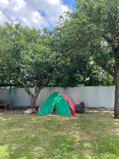 In the city of Лебяжье, Russia, a green and red tent is pitched on a patchy lawn in a backyard. The tent is set up under the canopy of a large, mature tree with dense green foliage, which casts dappled shadows on the grass. Behind the tent, a corrugated metal fence runs horizontally across the scene, serving as a boundary. To the left of the tent, a wooden picnic table and bench are visible. Adjacent to the tent, to the right, is a single, dark-colored car seat placed on the ground. The sky above is partly cloudy, with white clouds scattered across a blue expanse. The overall impression is one of a casual outdoor setting, possibly a campsite or a backyard play area.