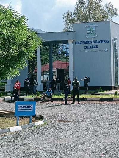 The image displays an outdoor daytime scene at Machakos Teachers College, located in Mumbuni North ward, Machakos County, Kenya. The central feature is a modern, multi-story building constructed of light-grey material, featuring a prominent glass-paneled entrance flanked by two white classical columns. The building's facade displays the college crest, "MACHAKOS TEACHERS COLLEGE," and "ISO 9001:2015 CERTIFIED."
Approximately 15-20 individuals are present in the scene, primarily adult males. They are observed standing or sitting in various small groups near the building's entrance and along the edge of a gravel-surfaced forecourt. Attire is casual, including an individual in a red "SUP" hoodie and another in a blue shirt seated on a low concrete barrier.
In the lower left foreground, a blue "PARKING" sign with a white right-pointing arrow is visible on a post within a grassy verge. The gravel forecourt exhibits several small puddles, suggesting recent moisture. Lush green foliage from trees borders the left side of the scene and is reflected in the building's glass entrance, alongside reflections of red-roofed structures. The sky overhead is partly cloudy, and distant hills or mountains are visible on the horizon in the background.