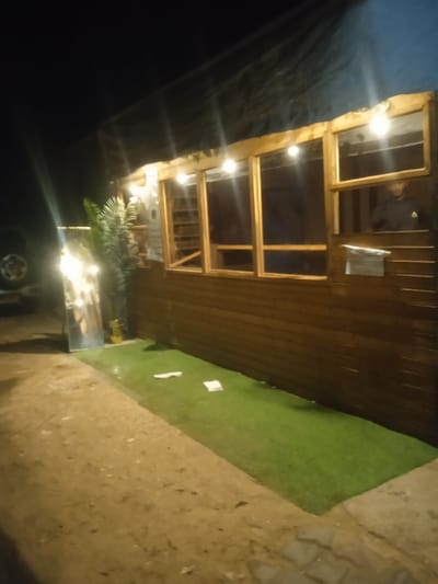 The image displays an outdoor night scene in Al-Mawasi, Palestinian Territories, featuring a well-lit, standalone structure. The structure is constructed with horizontal dark brown wooden planks and has a dark tarp-like material forming its sloped roof. Along the top edge of the wooden facade, multiple exposed incandescent-style light bulbs are strung, casting bright light on the immediate area.
The facade incorporates four window-like openings with light-colored wooden frames. The far-right opening functions as an open service hatch or window. Through this opening, a single individual is visible from the torso up, positioned inside the structure and looking outwards. This person wears a dark garment, possibly a hoodie, with a faint lighter-colored emblem or design on the chest area.
The ground in front of the structure consists of two distinct sections. Immediately adjacent to the wooden facade is a strip of green artificial turf, upon which several small, light-colored pieces of paper or debris are scattered. To the left of this turf, the ground transitions to an unpaved area of light brown dirt and gravel.
Standing upright on the green artificial turf, near the left side of the wooden structure, is a tall, slender, rectangular mirror or reflective panel, brightly reflecting the lights from the building. Beside this, a tall potted plant with prominent green leaves, contained within a gold-colored pot, is also positioned on the artificial turf. In the far left background, partially obscured by darkness, the front wheel and a portion of a dark-colored vehicle are discernible.