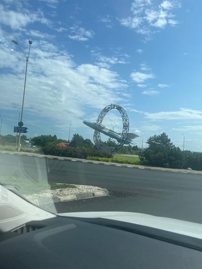 This media file shows an outdoor scene, likely in Bacău, Romania, on a clear, sunny day with scattered clouds. The primary object of focus is an airplane sculpture that appears to be suspended within a large, metallic loop structure. The airplane is painted in a grey and blue camouflage pattern with national insignia visible on its tail. This structure is situated near a road, with a white car visible in the foreground, suggesting the image was taken from inside a vehicle.

Behind the sculpture, a dense line of green trees forms a backdrop, with some red-tiled rooftops peeking through in the distance, hinting at a developed area. In the lower part of the image, there are streetlights and road signs. One sign with blue background and white text points to "SUCEAVA" and "VASLUI" with truck icons, indicating directions and possibly highway destinations. The road surface is asphalt, with a gravelly shoulder and a concrete curb.

The lighting suggests it is daytime, likely late morning or early afternoon, given the bright sunlight and clear sky. The overall impression is one of a roadside monument or landmark, possibly commemorating aviation history or serving as a local attraction. There are no people visible in the image. The scene conveys a sense of open space and a bright, pleasant atmosphere.