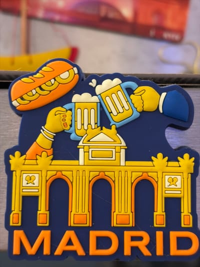 The image shows a dark blue magnet with a yellow and orange depiction of Madrid. At the top, an orange sandwich with yellow fillings is featured. Below the sandwich, two hands are shown raising mugs of beer. In the center, a yellow and white building resembling an archway is depicted. The word "MADRID" is written in large orange letters at the bottom of the magnet. The background is blurred and shows a hint of red and yellow objects, possibly part of a display or another magnet. There is no visible indication of Limnia, Northern Cyprus or Cyprus in the image.