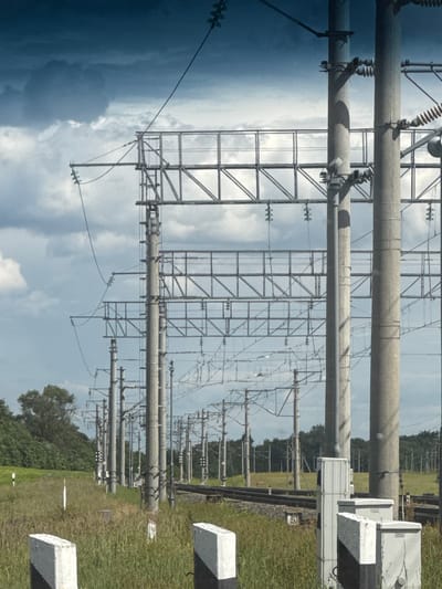 This is a landscape photograph taken outdoors, with the camera rotated 90 degrees counter-clockwise, so the image appears sideways. The primary subjects are a complex array of electrical transmission towers and equipment, characteristic of a power substation. Large, cylindrical grey conduits, likely high-voltage power lines, dominate the foreground, supported by tall metal lattice towers. Numerous smaller wires and insulators are visible, strung between the structures, creating a dense network against a cloudy sky. In the background, to the right, a grassy field stretches towards a treeline, with several concrete or stone markers, some with black and white stripes, placed along what appears to be a path or boundary. The weather appears overcast with dramatic, dark clouds on the left side of the image, contrasting with brighter, scattered clouds on the right. The overall impression is of a functional, industrial landscape under a dynamic sky, possibly a storm is approaching or passing. There is no visible text in the image.