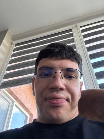 The image is a close-up, waist-up, selfie-style photograph of a man with dark hair and glasses. He is wearing a black t-shirt. The man is sticking his tongue out slightly. Behind him, a window with horizontal blinds is visible. To the left of the man, a window with a visible blue sky can be seen. The wall outside the window appears to be a peachy-orange color. A portion of what appears to be an animal's fur, possibly a dog or cat, is visible in the right side of the frame. The lighting suggests an indoor setting, likely during the daytime. The image does not provide any specific landmarks or clues that would indicate the location is in the city of Dibich, Bulgaria.