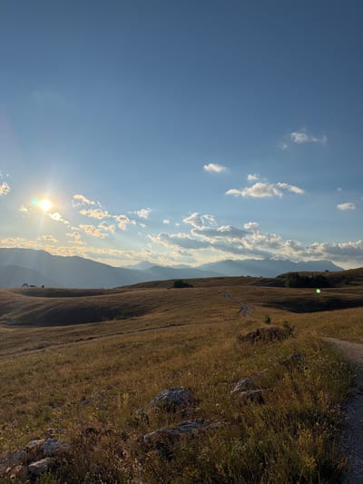 The image depicts a mountainous landscape in Dubljevići, Montenegro, bathed in the warm glow of the setting sun. The sun, a bright orb, is positioned low in the sky, casting long shadows and illuminating scattered clouds with golden light. The sky is a clear, vibrant blue, contrasting with the muted tones of the landscape.
The foreground is dominated by a dry, grassy hillside, dotted with small, purple wildflowers and rugged rocks. A narrow, winding dirt path snakes its way down the slope, suggesting a hiking trail. In the mid-ground, rolling hills and distant mountain ranges fade into a hazy silhouette, their forms softened by the atmospheric perspective.
The overall scene evokes a sense of tranquility and natural beauty. The time of day is clearly late afternoon or early evening, as indicated by the low sun and the warm lighting. The weather appears clear and fair, with only a few wispy clouds adorning the sky. There are no people or man-made objects visible besides the path, emphasizing the wild and remote character of the location. The image captures a moment of serene contemplation, inviting the viewer to appreciate the vastness and quiet majesty of the natural world.