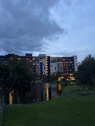 Captured at dusk, the scene depicts the Century City Hotel in Milnerton, South Africa. The multi-story building complex, illuminated with interior lights, stands alongside a body of water, possibly a canal or a section of a lake, reflecting the building's lights. Adjacent to the water, manicured green grass leads to a circular arrangement of what appear to be stone or concrete seating elements. The sky is overcast with dark clouds, and tree foliage is visible along the sides of the frame.