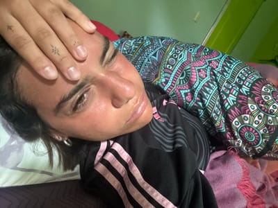 The image shows a woman lying on her back in bed in a room in Montañita, Ecuador, with coordinates -1.825 latitude and -80.752 longitude. The woman is wearing a black and pink Adidas shirt and a blue, pink, and gold patterned shirt. She has her eyes closed and her hand is resting on her forehead. Her face is slightly flushed. The background of the image shows a green wall and part of a pillow. There is also a pink and purple blanket visible in the bottom right corner of the image.