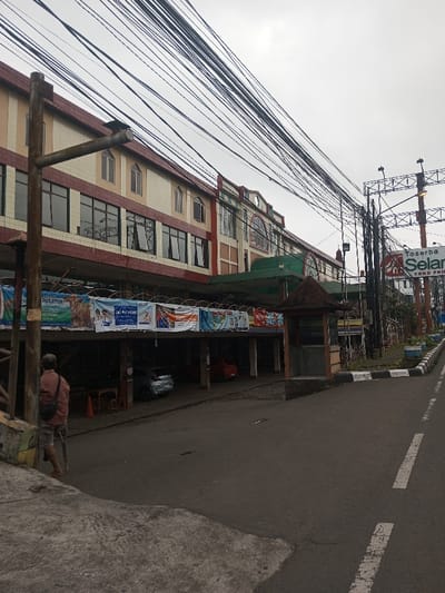 A street scene reveals a multi-story building adorned with advertisements. Below the building, cars are parked. Overhead, a web of electrical wires dominates the sky. A guard booth stands near the road, where painted lines demarcate lanes. A man walks along the side of the street. A large sign for "Toserba Selan" is visible in the background. The sky is overcast, suggesting a cloudy day.