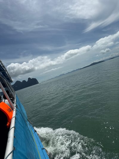 The image depicts a view from a moving boat, likely a tour boat, in the waters near Khlong Khian, Thailand. The boat's bright blue hull and white railing are visible on the left side of the frame, with an orange life vest attached to the railing. White wake churns from the boat's movement, indicating it is traveling at a moderate speed. The sea is a muted green color with small whitecaps and sunlit sparkles on the surface. In the distance, a cluster of dark, rugged islands with steep, rocky formations rises from the sea, characteristic of the limestone karsts found in Phang Nga Bay. The sky is partly cloudy, with dramatic formations of white and grey clouds contrasting against a light blue sky. The overall impression is one of travel and exploration in a scenic, tropical marine environment.