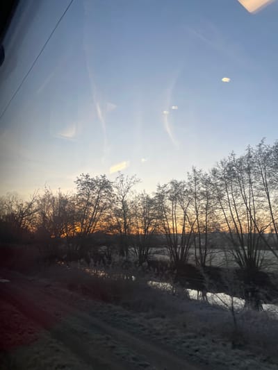 The image depicts a winter landscape seen through a window, possibly a train window, suggested by the internal reflection. Bare trees line a waterway, their silhouettes stark against the pale sky, tinged with the orange hues of sunrise or sunset. The ground is covered in frost. The reflections on the window create a soft, ethereal quality to the scene.