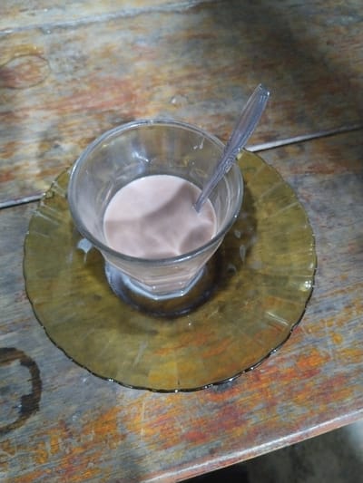 The image displays a clear glass containing a light brown or pinkish-brown liquid, likely a milky beverage such as chocolate milk or coffee with milk. A silver-colored spoon is immersed in the drink. The glass sits on a translucent, dark yellow or amber-colored glass saucer with a textured or scalloped edge.

Both the glass and saucer are placed on a rustic wooden table. The table surface is visibly worn, featuring distinct wood grain, various scratches, and areas of discoloration, including patches of gray, brown, and reddish-orange tones. There are noticeable liquid splatters on the saucer around the base of the glass. To the lower left of the saucer, a prominent dark, circular ring stain is visible on the wooden table, alongside other smaller liquid marks.

The lighting appears somewhat dim, suggesting an indoor setting or a low-light environment. The scene depicts a simple, casual moment, possibly a break for a drink. No people are visible, and there is no discernible text on any object. The location context provided for this image is Meunasahpantonlabu, Indonesia.
