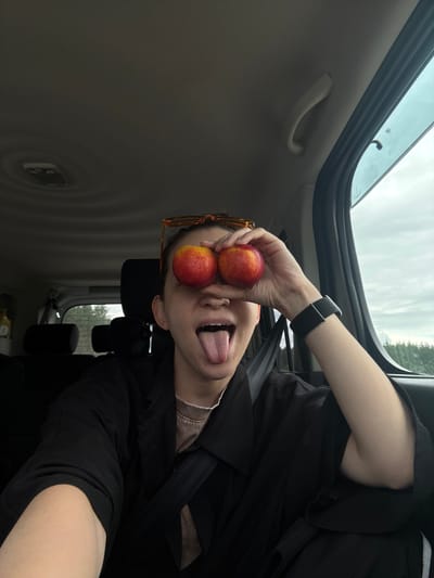 A person is in a vehicle, likely a car, with an open sunroof. The interior of the vehicle is light grey, with a grey handle visible near the roof. Outside the window, there is vegetation. The person, presumably the photographer, is holding two peaches over their eyes while sticking their tongue out. They are wearing an Apple Watch on their left wrist and a black shirt. A pair of orange-rimmed glasses rest on the side of their face. The location in the city of Baskan, Russia is unknown.