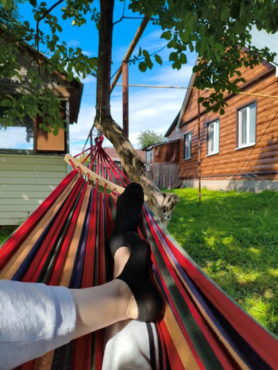 A person relaxes in a colorful hammock strung between two trees in a backyard. The hammock is made of red, blue, yellow, and green stripes. The person is wearing black socks and light-colored pants. The background features a wooden house with windows, green grass, and trees with lush leaves. The sky is bright blue with some white clouds, suggesting a sunny day. The scene evokes a feeling of leisure and relaxation. The location appears to be a rural or suburban backyard in Russia, possibly during the summer.