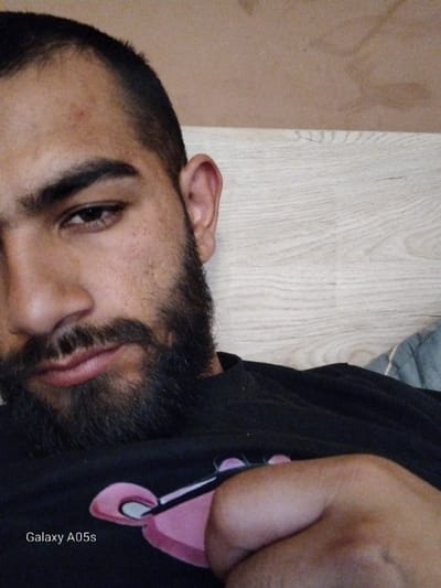 The scene is located in the city of Oreshene, Bulgaria. The image presents a close-up of a male subject, visible from the upper chest upwards, with his face turned towards the camera. He possesses short, dark hair, dark eyebrows, and a full dark beard and mustache. A small red lesion is present on his forehead. He wears a black t-shirt with a pink graphic design, identified as the Pink Panther, on the front. His right hand is partially visible, forming a fist and resting on his chest, partially obscuring the t-shirt's graphic. The background features a light brown wall or panel in the upper section and a light wood grain surface in the lower section, with a grey quilted textile partially visible in the bottom right corner. The watermark 'Galaxy A05s' is visible in the bottom left.