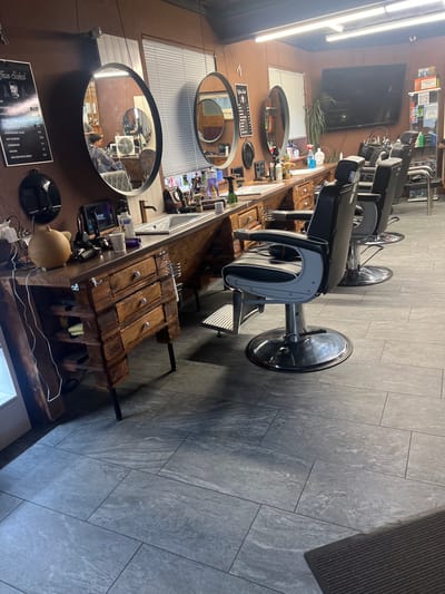Here is a detailed description of the image:
**Scene Overview:**
The image depicts the interior of a barber shop. The perspective is slightly angled, showing a row of barber chairs lined up along a wall with mirrors and workstations. The shop appears to be clean and well-maintained.
**People:**
A person is visible in the mirror of the workstation.
**Objects:**
* **Barber Chairs:** There are multiple black and gray barber chairs, with chrome bases.
* **Workstations:** Wooden workstations with sinks and drawers, appearing to be made from reclaimed wood.
* **Mirrors:** Oval mirrors mounted on the wall above the workstations.
* **Lighting:** Fluorescent lighting fixtures on the ceiling.
* **Flooring:** Gray tile flooring.
* **Wall Decor:** A menu board on the wall.
* **Miscellaneous Items:** Various barbering tools, products, and accessories on the workstations.
**Environment:**
* **Walls:** The walls are painted brown.
* **Windows:** Windows with white blinds are visible along the wall with mirrors.
* **Interior Design:** The shop has a rustic-modern aesthetic, with a focus on natural materials like wood.
**Location:**
Based on the menu board, it is determined that the location is in Liestal, Switzerland.