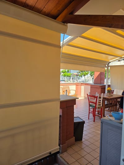 The image depicts a detailed view from inside a partially covered outdoor patio or terrace, looking outwards towards a street scene in Mosciano Sant'Angelo, Italy.

In the immediate foreground on the left, a large, light-beige or yellow retractable screen or wall blind extends upwards, occupying approximately one-third of the frame's width. This screen features two horizontal light-grey trim strips. At its base, a dark horizontal bar is visible, above reddish brickwork. On the far right foreground, a light-grey, vertically ribbed cabinet or storage unit is partially visible, topped with a white tray holding a blue bowl. The floor of the patio is covered with light-toned square tiles.

Above, a dark wooden beamed ceiling with light brown planks is visible on the upper left. Extending across the upper mid-ground is a bright yellow, multi-segmented retractable fabric awning, providing shade over the patio. White structural elements support this awning.

In the mid-ground, a reddish-brown brick counter or low wall structure is present on the left, next to which a dark-colored bin or bucket rests on the tiled floor. To the right of this, a wooden rectangular dining table is set with four chairs; one chair is distinctively red with a woven seat, another is white, and two others are partially visible. A small pot containing yellow flowers is placed on the dining table. Further left on the patio, beyond the brick counter and on a slightly lower level, two wooden slatted armchairs are visible. The patio's perimeter is defined by low reddish-brown brick walls and columns, with a section of textured light-colored wall in between.

Beyond the patio structure, a residential street is visible under a clear blue sky. A white wrought-iron fence runs along the street, which is lined with numerous green trees, some appearing to be palms or similar species. Portions of residential buildings are visible in the background, and a white car is parked on the street. No individuals are present in the scene, and no specific actions are depicted. The area is well-lit by natural sunlight, creating distinct shadows.
