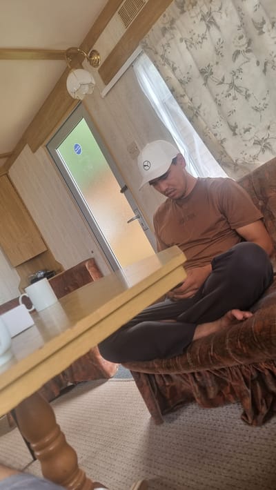 The image shows a person wearing a brown t-shirt and dark trousers, a white hard hat, and a face mask. The person is sitting on a sofa that is covered with a patterned brown blanket. The person appears to be in a room with light-colored walls and a patterned ceiling. There is a window with a view of greenery outside, and a light fixture on the wall. The overall setting appears to be a caravan or a similar mobile home.

The person is looking down towards a table in the foreground. There is a vent on the ceiling and a wooden cabinet. A red brick is visible near the window. The person's facial expression is not clearly visible due to the mask and angle, but their posture suggests they are engaged in some task or activity.

There is a circular sticker on the window with blue text and an icon, possibly indicating a warning or instruction. A white mug is also visible on the table. The lighting in the scene suggests it is daytime, but the specific time of day or weather conditions are not discernible from the image. The location context provided is Auchenblae, United Kingdom. No other text is clearly legible in the image.