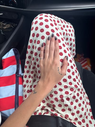 The image depicts a high-angle view inside the passenger compartment of a vehicle, likely a car. The primary subject is a person's left arm and hand resting on their lap. The individual's hand has long, almond-shaped fingernails painted with a dark grey or black gradient fading to a lighter tone near the cuticles. Visible tattoos on the hand include a thin band-like line on the ring finger, three small dots vertically aligned on the middle finger, a small series of curved lines on the outer palm near the pinky finger, and a faded, cursive word tattoo on the inner forearm near the wrist.

The person is wearing clothing that includes a black garment, possibly pants or a skirt, underneath a white fabric patterned with numerous red circular designs, each containing an internal star-like or flower-like motif, covering the thighs and lap. To the left of the legs, a portion of a red, white, and light blue striped zip-up jacket or similar garment is visible. The immediate environment includes dark car interior elements in the background, specifically parts of the dashboard or center console area with vents and controls in the upper left, and a dark seatbelt strap visible on the right. The specific location in the city of Texcalama, Mexico, cannot be determined from the visual information presented in this image.