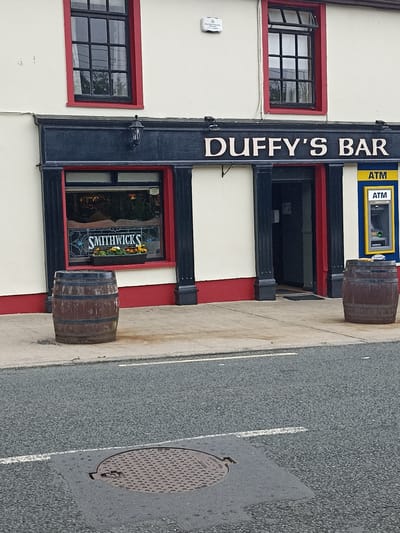 This image depicts the exterior of Duffy's Bar, located in Annagry, Ireland. The building features a cream-colored facade with red trim around its windows and base. Above the entrance, a dark blue sign reads "DUFFY'S BAR" in stylized, light-colored lettering. To the right of the entrance is a yellow ATM machine with "ATM" written in blue.
In front of the bar, to the left of the entrance, is a large wooden barrel. To the right of the entrance, another similar barrel is positioned. A window to the left of the entrance displays the word "SMITHWICKS" in stained glass and a flower box with orange and white flowers.
The ground in front of the building is paved, with a street in the foreground that has a manhole cover and white road markings. The sky appears overcast. There are no people visible in the image.