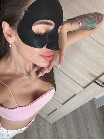 A female individual is depicted in an indoor setting within Krasnaya Zarya, Russia. She is reclined, with her eyes closed, wearing a black sheet facial mask that covers her forehead, eyes, and the bridge of her nose. Her exposed lower face displays freckles on the cheeks and around the mouth area. She has prominent, light pink lips. The individual is dressed in a light pink spaghetti strap top and white shorts or pajama bottoms featuring a subtle pink floral pattern and a small pink bow.
On her left ear, she wears a silver stud earring in the upper lobe and a gold-toned hoop earring with a single clear stone in the lower lobe. A silver chain necklace with a circular, clear-stoned pendant is visible on her chest. Her right arm, adorned with a colorful tattoo depicting floral elements and possibly a bird or dragon, is bent at the elbow, with her hand positioned near her right cheek. Her fingernails are long, almond-shaped, and finished with a light pink to white ombre manicure.
The immediate environment consists of a light-colored wooden floor with visible grain and a vertical, light wooden door featuring horizontal paneling and a silver-colored handle.