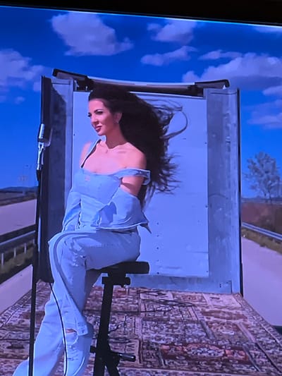 A woman is sitting on a stool inside the back of a semi-trailer truck. The truck bed is decorated with a Persian rug. The woman has long, dark hair blowing in the wind and is wearing a light blue off-the-shoulder top and matching pants. A microphone on a stand is to her left. The backdrop behind her is a projection of a bright blue sky with clouds. The truck is positioned on what appears to be a road or highway, with a landscape of trees and fields visible in the distance. The overall scene suggests an outdoor performance or photoshoot set within a truck.