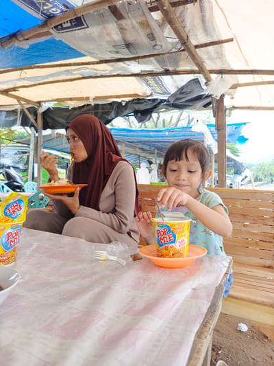 This image depicts an outdoor eating stall in Meurah, Indonesia, where an adult woman and a young girl are having a meal under a provisional canopy. The woman, wearing a dark reddish-brown hijab and a light brown long-sleeved outfit, sits cross-legged on a wooden bench behind a wooden table. She is eating noodles from an orange oval plate using a white plastic fork, with her mouth slightly open. To her right, a young girl, approximately 5-7 years old, dressed in a light blue patterned t-shirt, sits on a similar wooden bench. She is stirring a yellow 'Pop Mie' instant noodle cup (Chicken Curry flavor) with a small metal spoon, the cup resting on an orange plastic plate. Another identical, unopened 'Pop Mie' cup and a small white bowl are on the table to the left. The table surface is covered by a translucent plastic tablecloth featuring a repetitive light pink geometric pattern. The overhead shelter is a makeshift structure composed of wooden poles and beams supporting various materials, including blue tarps, white plastic sheets (some repurposed, showing partial text like 'GEL sehat' and 'TOP BRAND'), and black sheeting. In the blurred background on the left, elements like a black motorcycle, blue plastic chairs, and green foliage are visible. On the right, distant foliage, possibly palm trees, and faint figures in light clothing suggest an open area or the coast beyond the stall. The ground surrounding the table is exposed dirt.