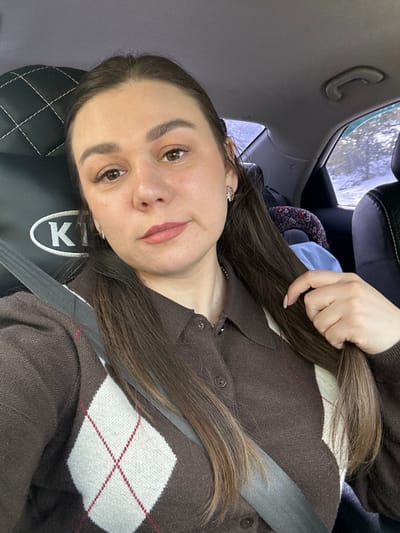 This image depicts a female subject seated inside a vehicle. The scene is identified as being in Krasnye Gorki, Russia. The subject, a woman with long, dark brown hair parted to her left, is looking directly forward towards the camera. She has light-colored eyes, defined eyebrows, and appears to be wearing subtle makeup, including a light pink lip color. A small, ornate earring is visible in her right ear. She wears a brown collared cardigan featuring a white and maroon argyle pattern on the front, with buttons running down the center. A silver-toned chain necklace is visible around her neck. A gray vehicle seatbelt diagonally crosses her chest. Her right hand is shown lightly grasping a section of her hair near her shoulder.
The immediate environment is the interior of a car. A black, quilted headrest with a white "KIA" logo is partially visible behind her left shoulder. In the background, beyond the subject, the vehicle's rear interior shows parts of a dark seat, a blue object, and a patterned child car seat. Through the rear and side windows, a distinct snowy landscape is visible, characterized by snow-covered ground and dark, bare trees, indicative of a winter outdoor environment. The car's ceiling and a grab handle are also partially visible.
