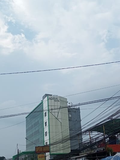A tall, modern building with the sign "AGRIA HOTEL" stands against a cloudy sky. The building features a green-tinted glass facade on one side and a white-and-gray side. Overhead power lines crisscross the foreground, obscuring the view of the structures below. A sign reading "OPTIK TAJUL" is visible, advertising "SOFLENS" for "50.000".