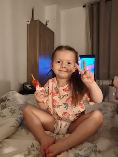 A young girl with brown hair is sitting cross-legged on a bed. She is wearing a pink t-shirt with a strawberry pattern and a diaper. In her right hand, she holds a popsicle. She is smiling and making a peace sign with her left hand. The background shows a wardrobe, a television screen with colorful content, and curtains. The scene appears to be indoors, likely a bedroom, during the evening or nighttime, as indicated by the artificial lighting and the dim ambiance. The girl's expression conveys happiness and playfulness. The t-shirt has visible text that reads "STRAWBERRY."