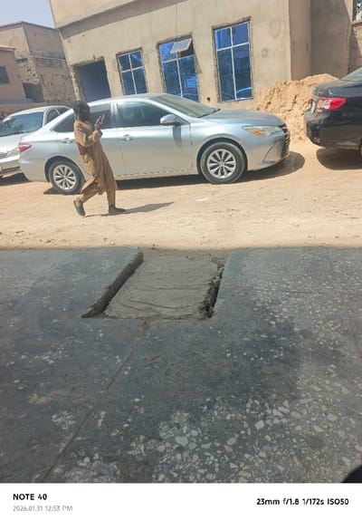 The image depicts an outdoor urban or semi-urban scene in Kofar Ruwa, Nigeria, under direct sunlight at midday (12:53 PM).
In the foreground, a dark, speckled concrete or terrazzo surface covers the lower half of the frame. This surface contains an irregular, linear trench approximately 15-20 cm wide, filled with lighter-colored dirt or sand, forming a rough L-shape that extends from the left edge towards the center.
The midground features an unpaved, light brown dirt road or pathway. A male individual, dressed in a loose-fitting light brown garment and dark shoes, walks from left to right across this pathway. He holds a dark object, consistent with a mobile phone, up to his face with his right hand, appearing engaged with the device. A distinct shadow extends from his body onto the dirt ground. Three vehicles are parked on this unpaved surface: a silver Toyota Camry sedan is prominent in the center, facing right; a portion of another silver sedan is visible behind it to the left; and the rear of a black sedan is visible to the right.
The background includes a two-story building with a light brown, possibly unfinished stucco or concrete facade. The upper floor features three rectangular windows with reflective blue glass, one of which appears partially open. To the right of the building, a substantial pile of light brown sand is present. Additional building structures are partially visible on the far left.
