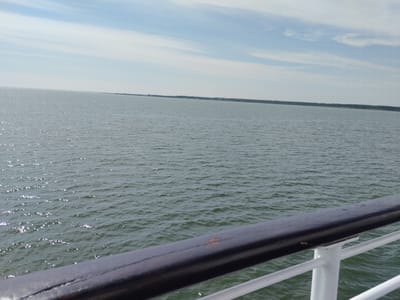 The image shows a wide expanse of calm, green water under a bright, hazy sky with scattered white clouds. In the foreground, a dark wooden railing and white metal supports of a ship's deck are visible, angled across the bottom right of the frame. The distant horizon line is sharply defined, separating the sea from a faint, dark strip of land that appears to be lined with trees and possibly some structures. This landmass stretches across the middle ground of the image. The overall scene evokes a sense of peaceful travel over water, likely on a ferry or cruise ship, with the coast of Kuivastu, Estonia, in the distance.