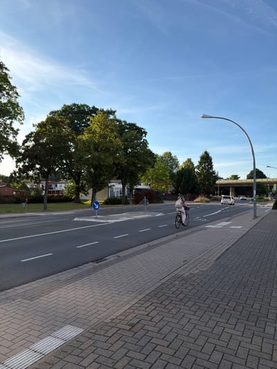 The image depicts a street scene in Nienburg/Weser, Germany, under a clear blue sky with wispy clouds. A woman is cycling on the right side of a road, wearing light-colored clothing and a backpack. The road has lane markings and a designated bicycle lane. Adjacent to the road is a sidewalk paved with grey and brown bricks, featuring tactile paving for visually impaired pedestrians. Trees line the left side of the road, with buildings visible in the background. Further down the road, a white car is driving, and an overpass or bridge is present in the distance. A street light arches over the scene.