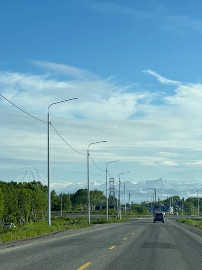 The image depicts a road in the city of Svetly, Russia, under a partly cloudy blue sky. The road, which is asphalted and has yellow lane markings, stretches into the distance. Several streetlights line the right side of the road, with power lines running between them. On the left, a dense line of green trees forms a backdrop, and beyond the trees, snow-capped mountains are visible. A blue SUV is driving away from the camera on the road. In the middle ground, there are various structures, including what appear to be buildings and power transmission towers. The overall scene suggests a semi-rural or suburban environment, possibly on the outskirts of the city, with a dramatic mountainous landscape in the background.