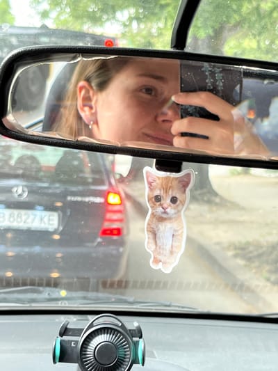 A selfie is taken in the rearview mirror of a car, showing a person holding a phone and a fluffy orange kitten keychain hanging from the mirror. The car is in traffic in Orizari, Bulgaria, evidenced by the license plate "B 8827 KE." The image is a close-up, with the driver's face and the kitten keychain as the primary subjects. The background shows other cars and trees, suggesting an outdoor setting.