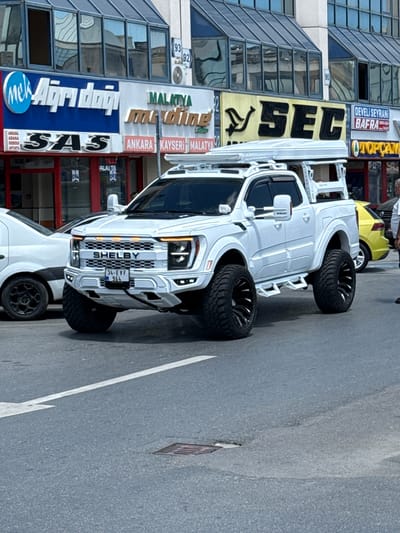 The image shows a white Shelby truck parked on a city street in Bayrampaşa, Turkey. The truck has oversized tires and a front grill with "SHELBY" displayed prominently. The license plate reads "34-EVF 964". In the background are several business signs, including "Airdağ", "SSYASS", "mayding", "SEÇ", "DEVELİ SEYRANI", and "BAFRA". The street appears to be paved, and there is a white lane marker. The buildings are modern and made of glass.