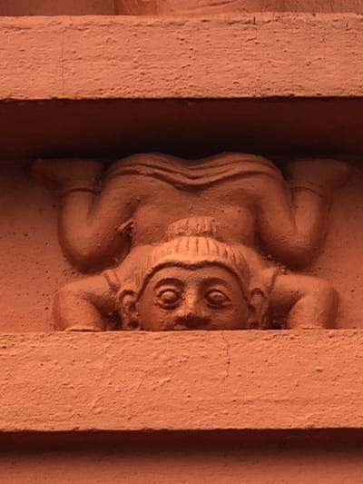 The image shows a terracotta bas-relief sculpture of a figure in a contorted pose, possibly performing yoga or acrobatics. The figure's head is at the bottom, with its face clearly visible. It has large eyes and a headdress. The body is bent backward, with legs in the air and arms supporting the weight. The sculpture is set into an architectural element, likely a wall or facade, made of a similar terracotta material. This sculpture is located on the outer wall of the Vidyalankara Pirivena in Peliyagoda, which is within the administrative limits of Sri Jayewardenepura Kotte, Sri Lanka. The lighting suggests it is outdoors during daylight.