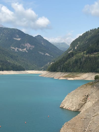 The image depicts a scenic view of a mountain lake in the Italian Alps, specifically the artificial lake of Sauris (also known as Lago di Sauris) near the village of Lateis (Latais). The lake's vibrant turquoise water occupies the foreground and midground, with visible layers of exposed earth and rock along its shoreline, indicating a low water level. Several small orange buoys are scattered across the water's surface, marking a boundary or hazard.
In the background, steep, densely forested mountains rise on either side of the valley. The slopes are covered in a mix of evergreen and deciduous trees, with patches of exposed rock and cliffs visible. The sky above is a clear blue, with scattered white clouds. The overall impression is one of natural beauty and dramatic mountainous terrain, characteristic of the Carnic Alps.