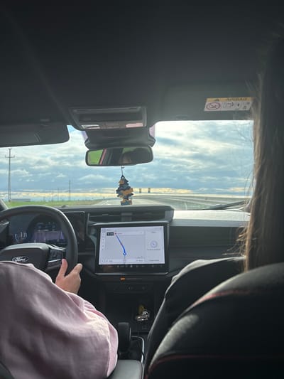 The image depicts the interior of a Ford vehicle from a rear passenger's perspective, looking forward on a multi-lane highway identified by the navigation system as DN56, in the general vicinity of Moțăței, Romania. The time is 16:23, and the ambient temperature is 11° Celsius.
A driver, wearing a light pink long-sleeved garment, has their left hand positioned on the black leather-wrapped steering wheel, which features a Ford logo and integrated controls. The digital instrument cluster displays a speed of 108.7 (likely km/h), "FM 90.6", and other vehicle data. A large central infotainment screen is active, showing a navigation map with a blue route line, indicating a "2nd exit" 14 km ahead and 136 km remaining to the destination. The phone function on the screen indicates "No device connected." Below the central screen, a gear selector and a small collection of keys are visible.
To the right, the back of a passenger's head, with long dark hair, and their dark-colored shoulder are visible. The passenger seat features red stitching. A "Little Tree" air freshener, colored yellow, green, and brown, hangs from the rearview mirror. An "AIRBAG" warning decal is affixed to the passenger-side sun visor.
The exterior scene through the windshield shows a straight, paved highway with a visible median strip. The horizon is flat, characteristic of a rural or semi-rural landscape, with several utility poles or power line towers positioned along both sides of the road. The sky is partially cloudy with significant patches of blue, suggesting fair weather. Distant trees and what appear to be industrial or agricultural structures are visible on the horizon.