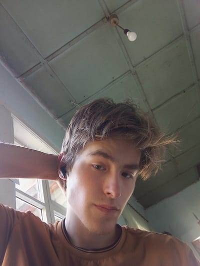 A young man with tousled brown hair is positioned in the foreground, looking directly at the camera. He has light skin and is wearing a brown crew-neck t-shirt. One arm is raised, with his hand resting behind his head. He appears to be wearing a black wireless earbud in his visible ear. The background consists of a light-colored ceiling with visible beams and a single lightbulb hanging from a cord. To the left, a window with a grid-like frame is partially visible, suggesting an indoor setting. The overall lighting suggests it is daytime. There is no indication of the specific location within Зертас ауылдық округі, Kazakhstan.