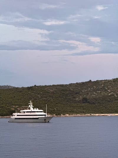 A large, white motor yacht with multiple decks and a sleek design is anchored in the calm, blue waters of the Adriatic Sea. The yacht is positioned horizontally across the frame, with its bow pointing to the right. Behind the yacht, a densely forested, green hillside slopes down towards the water's edge. The sky above is overcast with soft, pale blue and grey clouds. The scene appears to be a serene coastal landscape, possibly near the city of Primošten, Croatia, given its characteristic coastline and vegetation. There are no people visible in the image. The overall impression is one of tranquility and a luxurious maritime setting.