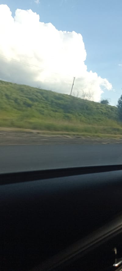 The image is a vertical shot taken from inside a vehicle looking out towards a cloudy sky and a grassy hillside. In the foreground, the dark interior of the car is visible, likely the dashboard or door panel. A grey road is visible in the middle ground, with a blur indicating motion. The background shows a green, grassy hillside with a few sparse trees and what appears to be a telephone pole. The sky is predominantly blue with a large, bright white cloud on the left side of the frame. The photo was taken in Valky, Ukraine.
