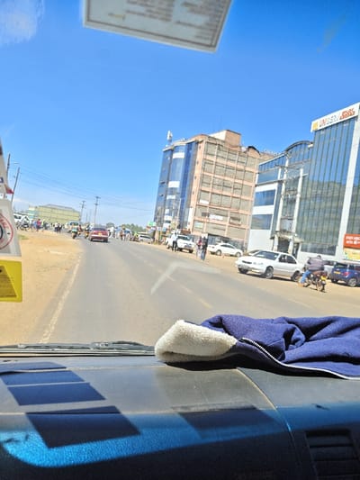 The image depicts a daytime street scene in Kapsabet, Kenya, viewed from the interior of a vehicle. A folded blue fleece-lined garment with a white inner lining rests on a dark grey dashboard in the lower right foreground. Rectangular shadows are cast on the lower dashboard by sunlight. A blurred, light-colored rectangular object is partially visible hanging from the vehicle's interior at the top center, and a yellow object with a partial white sticker reading "CARE" is visible on the left side of the windshield.
The main subject is a two-lane paved road with dirt shoulders, extending into the distance under a clear blue sky. On the road and shoulders, multiple vehicles are present, including a red sedan, a white sedan, a blue car, and several motorcycles (boda-bodas), some carrying riders and others parked. Pedestrians are scattered along both sides of the road and in the middle distance. Utility poles with power lines line the left side of the road.
On the right side of the road, a cluster of multi-story commercial buildings is visible. The prominent building closest to the viewer features a blue glass facade with "UNISERV" and "STREET" signage at its upper level. Adjacent to it stands a multi-story building with a light brown, concrete-gridded facade. Further along, another multi-story building with a blue glass facade is present. In the far distance on the left, additional buildings are discernible.