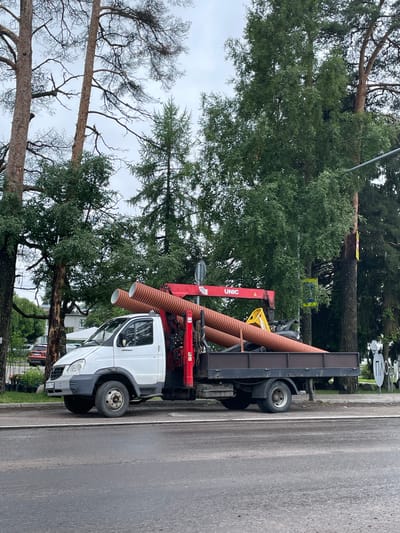 A white truck with a red UNIC crane is parked on a street in the city of Plodovoe, Russia. The truck is carrying two large, orange corrugated pipes. The truck is parked on the right side of the road, with a sidewalk and trees behind it. The sky is overcast, and there are some buildings visible in the background. The scene appears to be a construction or utility work site.