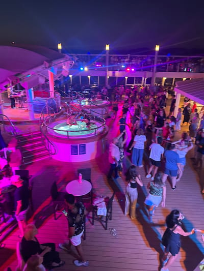 The image depicts an outdoor party scene at night, possibly on a cruise ship deck or a waterfront venue. The area is illuminated by vibrant purple and pink lights, with some blue and yellow lights also visible. In the background, a DJ is seen playing music on a keyboard setup.
The main focus of the image is a large gathering of people dancing and socializing on a wooden deck. Several individuals are actively dancing, with others conversing and standing around. There are also two circular hot tubs in the mid-ground, with people partially submerged in the water.
In the foreground, some people are seated at small tables, and one person is looking at their phone. The overall atmosphere appears festive and energetic.
Regarding the location in Salobreña, Spain, the image itself does not contain any specific visual cues that would allow for definitive identification of Salobreña. The scene is generic to many open-air entertainment venues, particularly those found on cruise ships or in tourist resort areas.