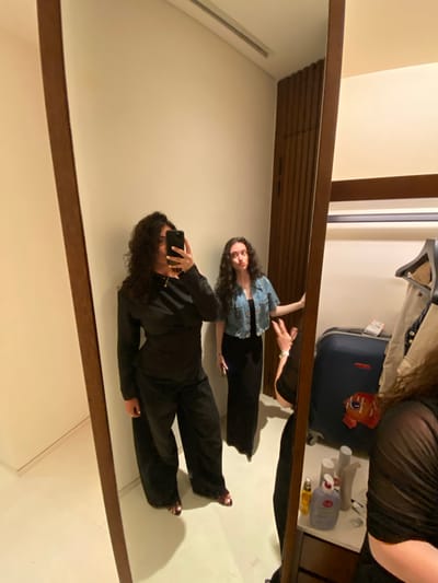 A photo of two women reflected in a large, rectangular mirror with a brown frame, likely taken in a hotel room in El Caracol, Mexico. The room contains a dark blue suitcase, a clothing rack with items hanging, and toiletries on a surface. One woman in the mirror, wearing black pants and a jean jacket, is standing and making a gesture with her hand. The other woman is wearing all black and is taking a photo with a mobile phone.