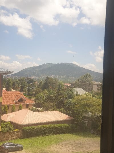This image, captured from an elevated perspective in Mumbuni North ward, Kenya, presents a residential landscape under a partly cloudy sky. In the foreground, a dark vertical window frame partially obscures the right edge. A green, unpaved area or lawn occupies the lower portion, featuring visible tracks. A dark grey four-door hatchback car is parked on the left side of this grassy expanse. Behind the lawn, a low, dense green hedge separates it from a single-story building with a light brown corrugated metal roof and light-colored walls. To the left of this building, a partial stone wall is visible. To the right, a solitary individual in light clothing is seated or kneeling amidst green foliage. Further back, numerous residential houses are visible, characterized by reddish-orange terracotta tile roofs and other lighter-colored roofs, interspersed among dense green trees. The most prominent house on the left features a distinct terracotta tile roof and light walls with a gable. The background is dominated by a large, green, tree-covered mountain or hill, showing scattered signs of urban development on its slopes. The sky above is light blue with scattered white cumulus clouds, indicating daytime conditions.