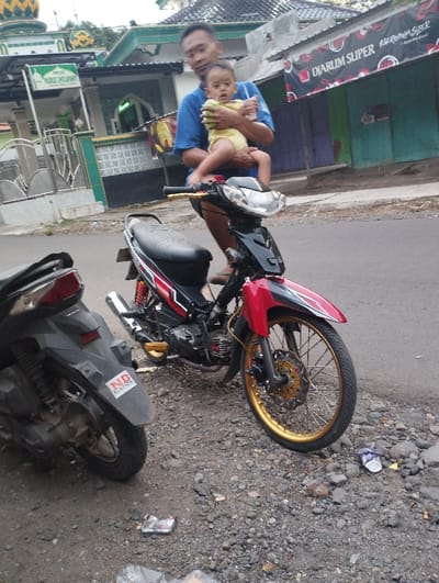 The image captures a man holding a baby, positioned on a red and black motorcycle, parked on a gravel and dirt roadside. The man, wearing a blue t-shirt and shorts, gently holds the baby, who is dressed in a yellow romper and appears to be sitting on the motorcycle's front. Both are looking towards the camera.
The prominent motorcycle is a red and black underbone style, featuring distinctive gold-colored rims, footpegs, and a front brake disc. Partially visible on the left is another dark-colored scooter, whose rear fender has a sticker that reads "ND HONDA". The ground is rugged, consisting of gravel and dirt, with some discarded plastic wrappers and other debris.
In the background, on the left side, there's a building with a green roof and a golden dome, characteristic of a mosque or prayer house. A green sign clearly states "MASJID NURUS SHOUFWANT". To the right, a shop is visible with a red banner across its front, displaying "DIARUM SUPER" and "Djarum Super Mld". The buildings are topped with corrugated metal roofs.
The scene unfolds during the daytime, under soft, diffused lighting, suggesting an overcast sky or a time of day with indirect sunlight. The setting evokes an everyday street or neighborhood scene in a Southeast Asian country, aligning with the location context of Kecamatan Panji, Indonesia.