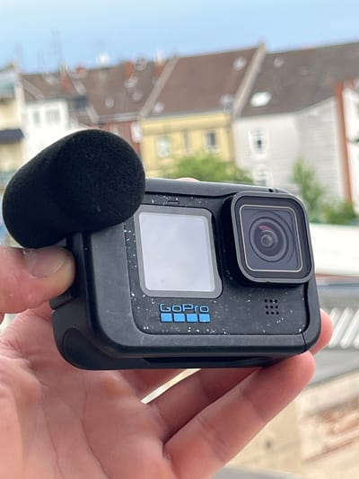 A close-up, outdoor, eye-level shot features a hand holding a black GoPro action camera with a black foam wind-muffling microphone attached to the side. The camera has a small screen on the front, a lens with a circular design, and the GoPro logo in blue. The camera is speckled with tiny white dots. The background is blurred, showing residential buildings with red-tiled roofs, windows, and some green foliage, suggesting a city environment. The sky is a pale blue. The scene implies the camera is being used for recording or photography. No specific landmarks or street names are visible to pinpoint a precise location within Neuss, Germany.