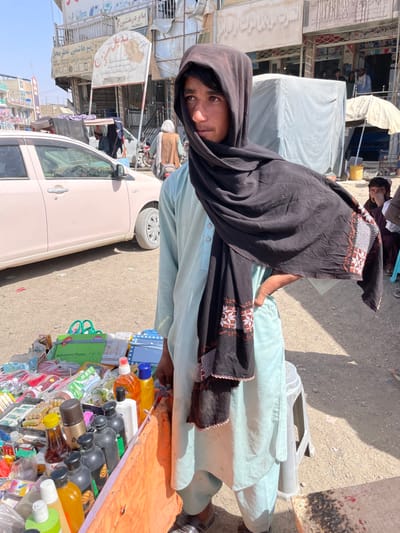 This image captures a street market scene in Spin Boldak, Afghanistan, under bright daylight.
A young male individual, approximately late teens to early twenties, stands slightly off-center to the right, facing towards the left side of the frame with his gaze directed outwards. He wears a light teal or pale blue long-sleeved tunic (likely a shalwar kameez top) with a buttoned collar. A dark brown or black shawl is draped over his head and shoulders, with an embellished section featuring white and reddish-brown geometric embroidery visible on the portion of the fabric flowing outwards to his right. His right hand is placed on his hip. He wears dark sandals.
Immediately to the left of the individual is a makeshift market stall, its front covered by an orange-brown fabric. The stall displays various merchandise, including multiple bottles of liquids (shampoo, oils, or similar toiletries) in assorted colors (black, white, yellow, clear, brown), an aerosol can, small plastic containers, and packaged goods. A green package labeled "Belladonna Plaster" is clearly visible, along with other items suggestive of general household or personal care products.
In the mid-ground, a light pink or beige four-door sedan is parked facing towards the background on the dusty, unpaved street. Behind the car, further street activity is visible, including other pedestrians and motorcycles. The background features multi-story commercial buildings housing shops, identified by signs with local script, one of which visibly includes the English word "PHARMACY" and another potentially "Mobile Sales." Further to the right in the background, another individual is seated under a white market umbrella. The sky is clear and light blue, indicating fair weather. The ground is consistently unpaved and dusty throughout the scene.