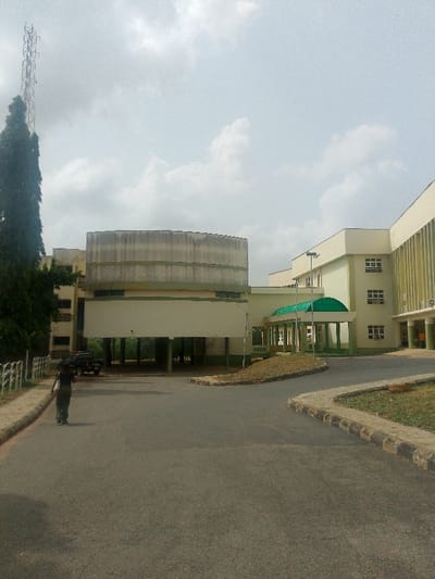 An outdoor scene in Adzho, Nigeria, displays an institutional complex under an overcast sky. In the foreground, a dark-paved road curves right, bordered by concrete curbs and grassy verges. A lone individual, dressed in dark clothing, walks away from the camera on the left side of the road. To the left, a tall, slender tree stands prominently, with a metal lattice tower partially visible behind it. Multiple multi-story buildings dominate the midground. The leftmost building features a distinctive light-colored, circular upper section with darker banding, and an elevated ground floor creating an open underpass where a dark-colored vehicle is parked. A second, lighter-colored building to the right exhibits vertical green accents on its facade and multiple windows. A connecting structure between the buildings features a prominent green arched canopy over an entrance. White metal railings line the left side of the road, and a streetlight pole stands near the center.